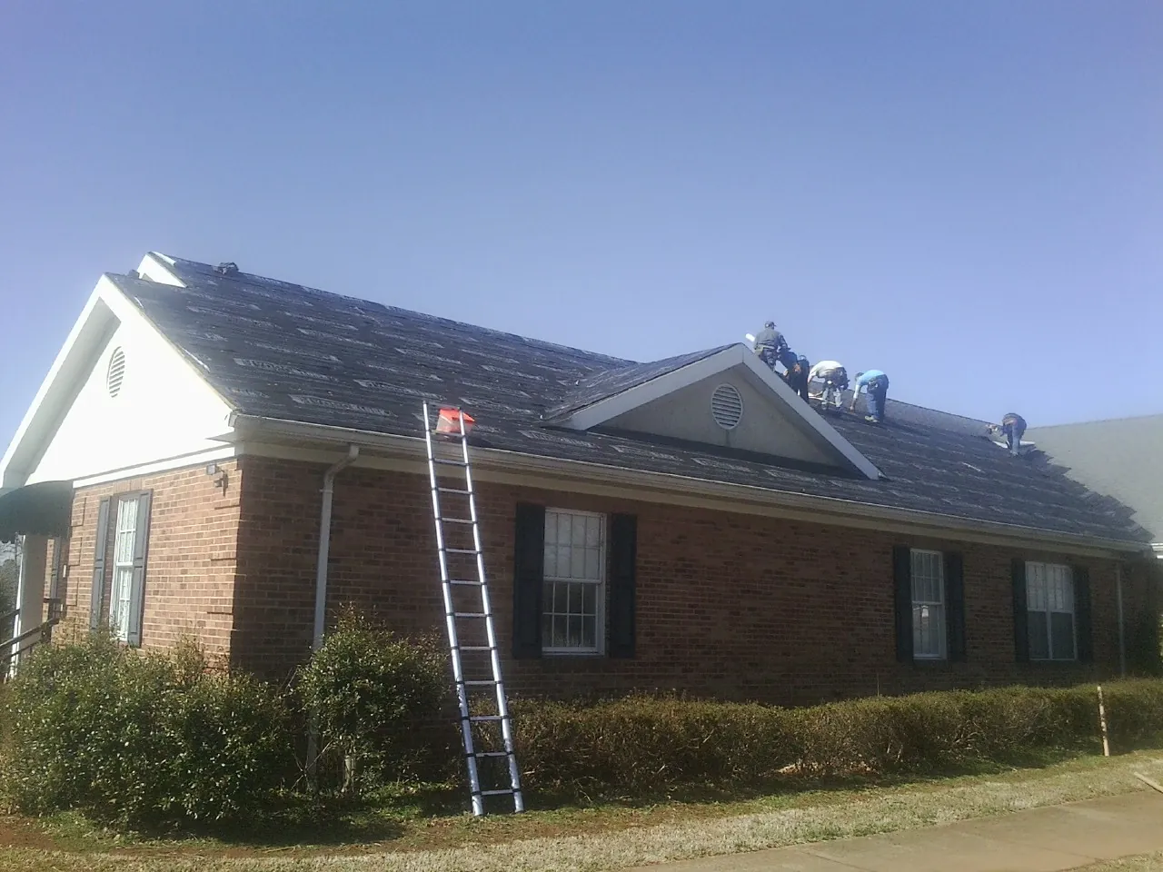 Crew tearing shingles off a brick ranch roof with a ladder set — Emergency Roof Repair work by our crew in Basile